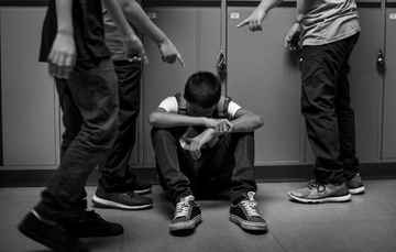 Black-and-white photograph of a seated student being surrounded and pointed at by other students in a school corridor.
